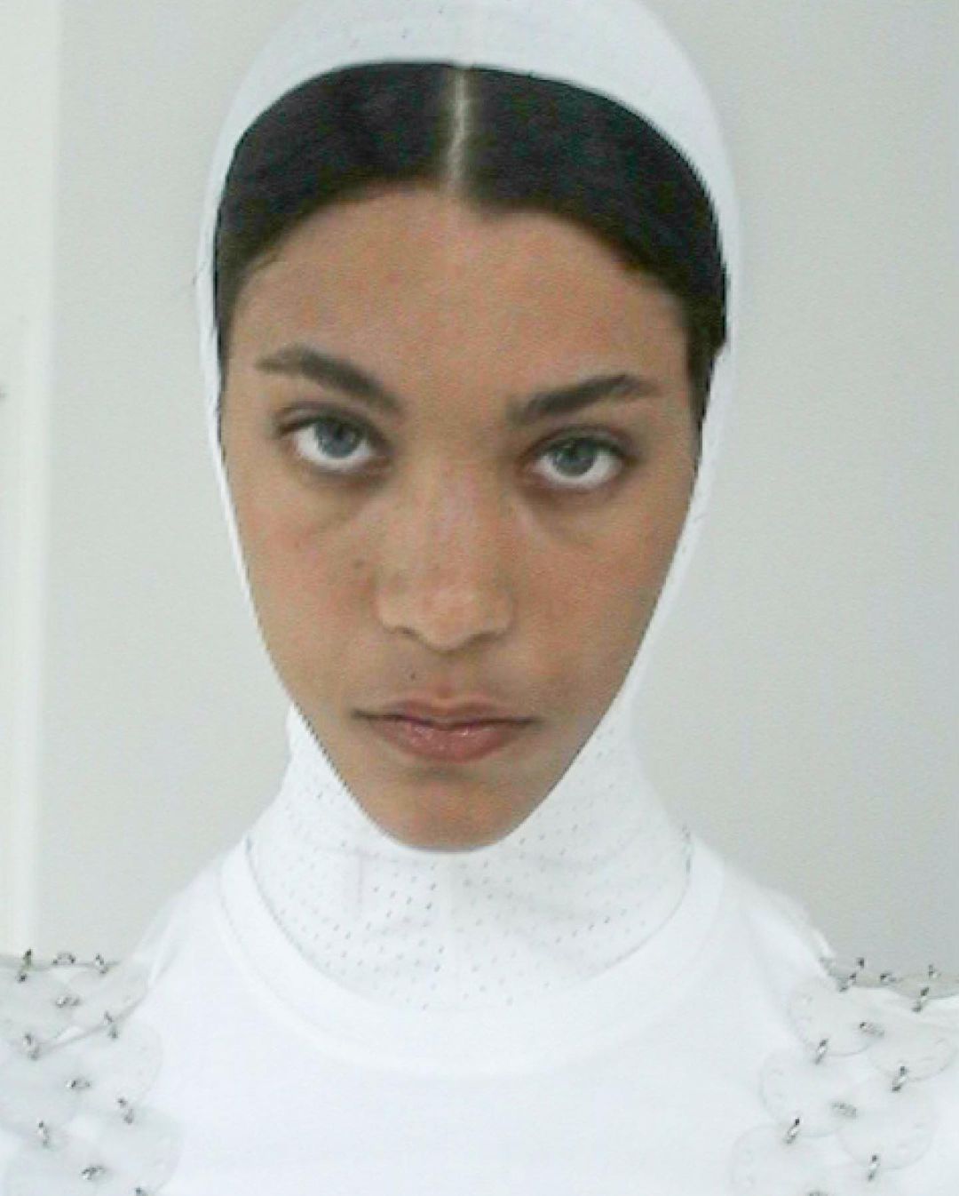Close-up portrait of a woman with fair skin, wearing a white head covering and top. The image is brightly lit with a plain background.