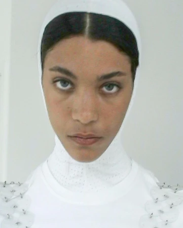 Close-up portrait of a woman with fair skin, wearing a white head covering and top. The image is brightly lit with a plain background.