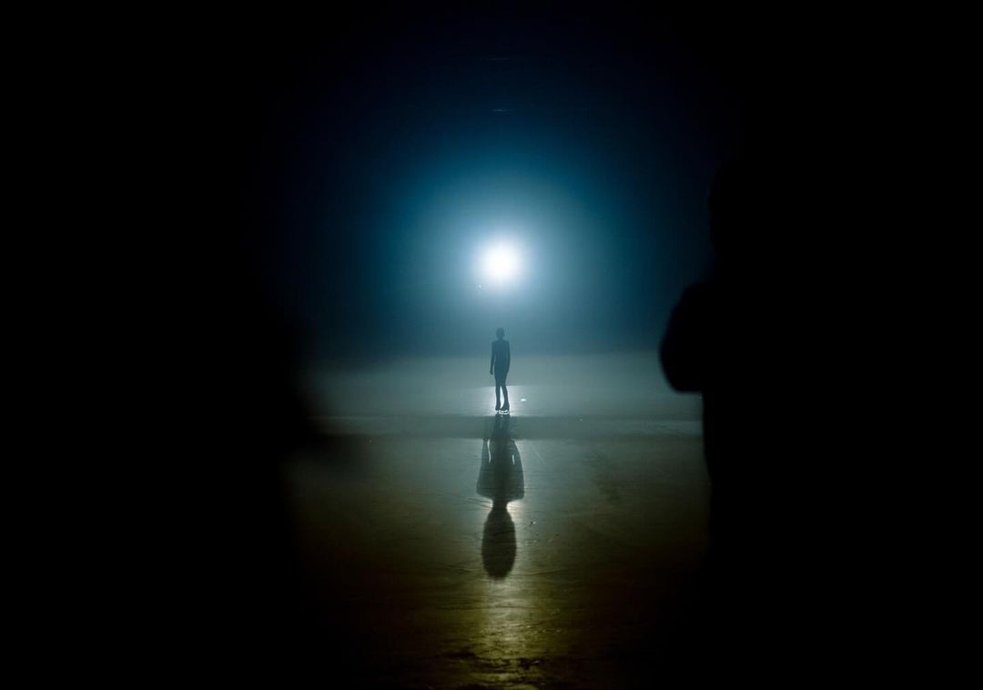 Lone figure stands beneath bright light in a dark, expansive space, casting a reflection on a reflective surface; silhouettes in foreground.