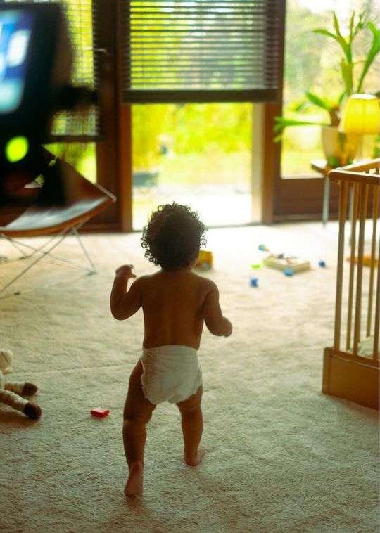 Infant explores sunlit interior, walking on carpet toward open doors, toys scattered nearby, and a crib in the background.