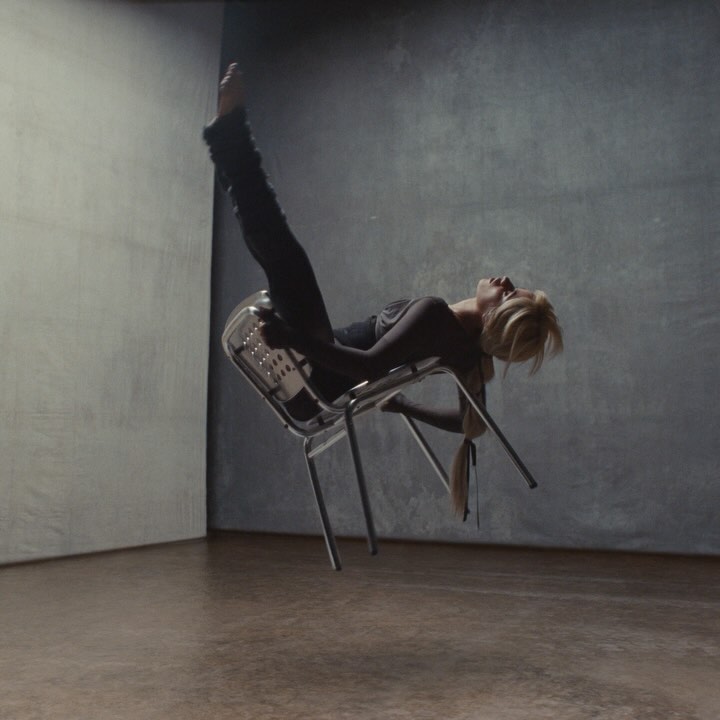 A woman with blonde hair balances upside down on a chair in a studio with a neutral grey background, showcasing an abstract artistic pose.