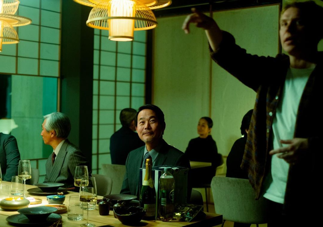 A group of people gathered around a table in a warmly lit room, enjoying a meal, with an Asian businessman at the center.