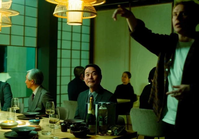 A group of people gathered around a table in a warmly lit room, enjoying a meal, with an Asian businessman at the center.