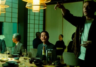 Group of people seated at a dining table with a standing man gesturing above them in a warmly lit restaurant interior