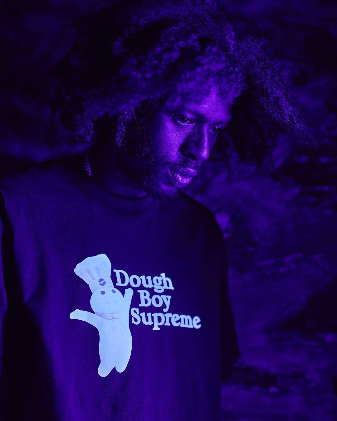 Portrait of an adult male wearing a black t-shirt with a Dough Boy logo in a purple, dimly lit environment. The subject looks downward.