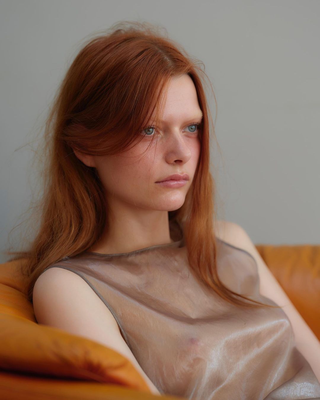 Portrait of a young, fair-skinned adult female with long red hair in a sheer top, reclining on an orange couch against a grey background.