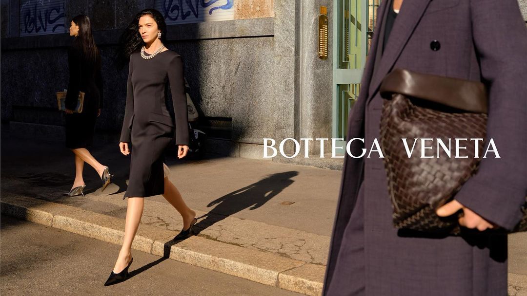 Fashion ad features two adult women in black dresses walking on a city street carrying a brown woven leather bag by Bottega Veneta.