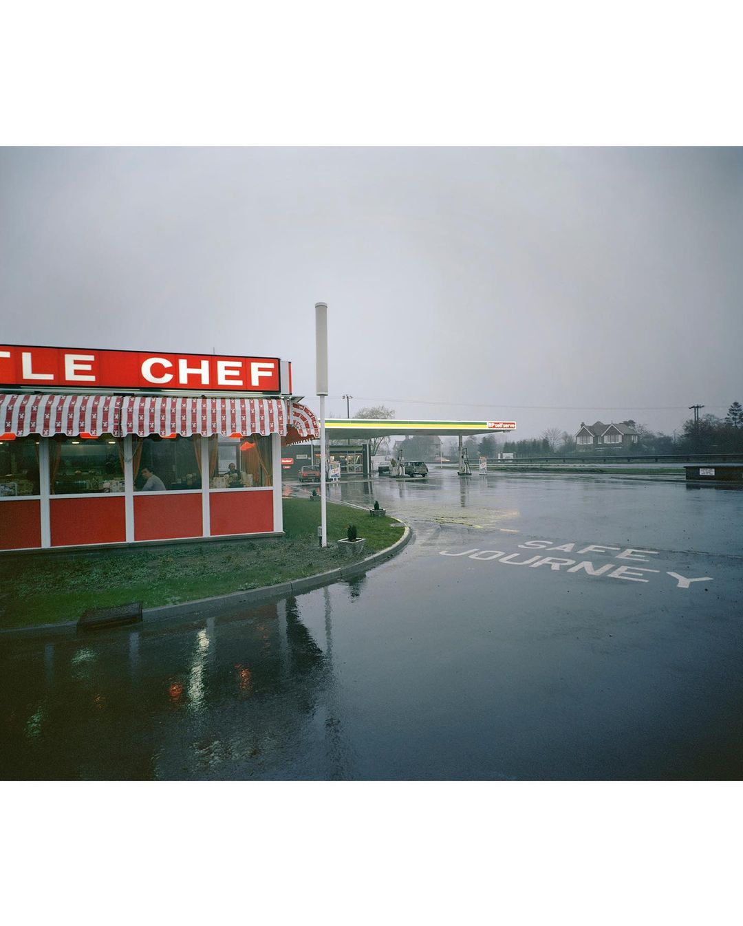 A roadside Little Chef restaurant and petrol station on a rainy day, reflecting the overcast sky and wet asphalt in the foreground.