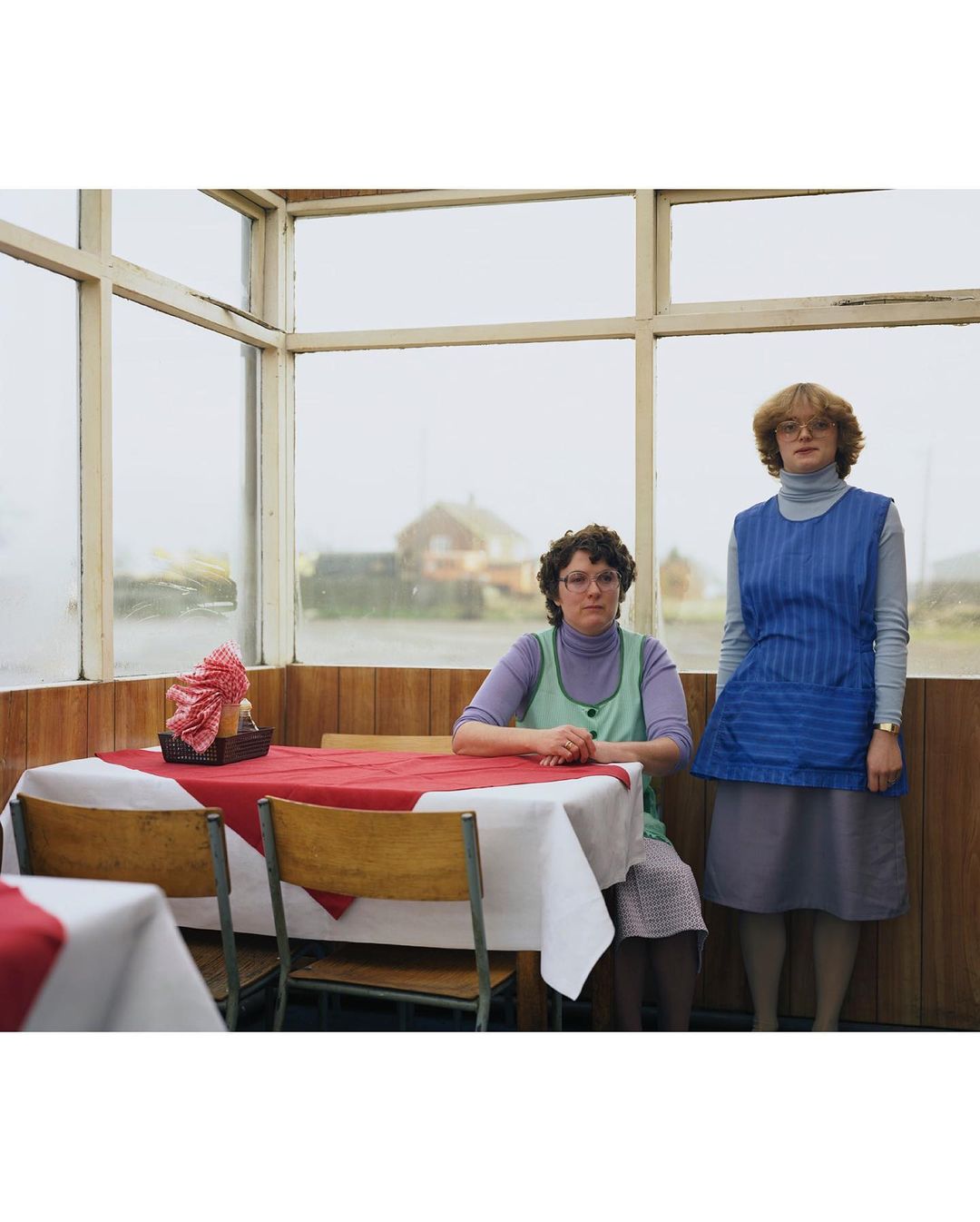 Two adult women in matching waitress uniforms stand indoors near tables set with red cloths, in a brightly lit eatery with large windows.