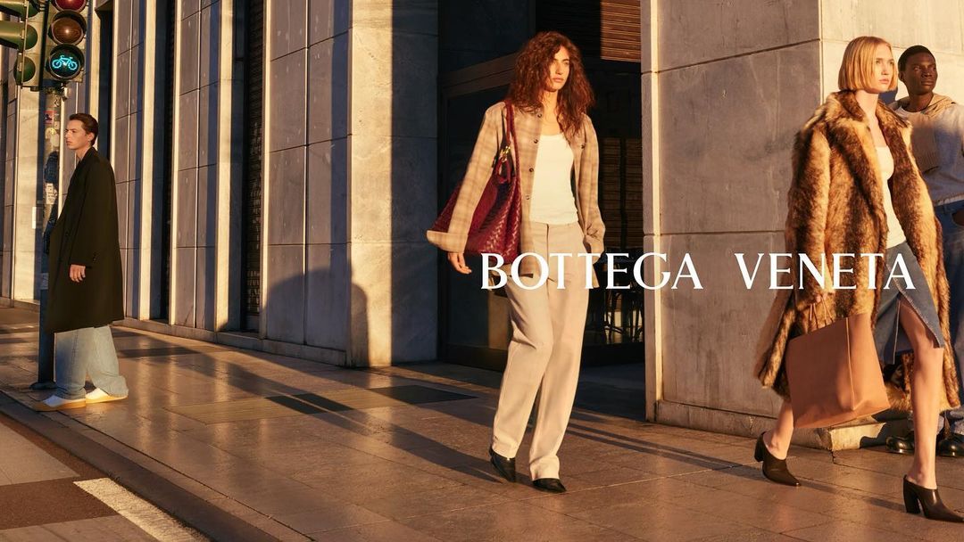 Fashion editorial shot on city street features two stylish adult females in coats, one male, walking near architecture with hard shadows.