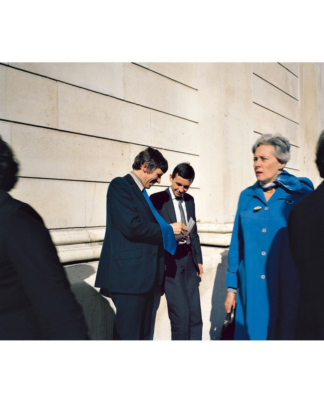Candid outdoor shot of two men in suits looking at a card and a woman in a blue coat outdoors in front of a light-colored wall.