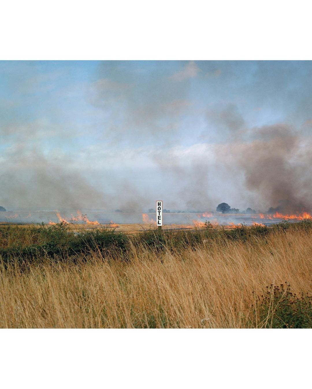 Rural landscape shows a field of golden grass and a distant fire, marked by a HOTEL sign, under a blue sky filled with smoke.