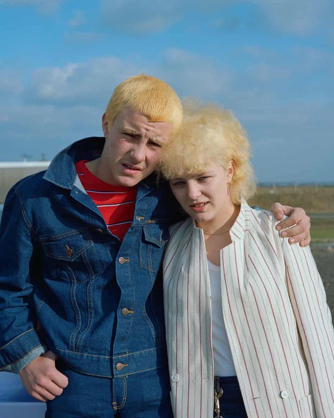 A blonde-haired male and female stand together, embracing in an outdoor setting with a blue sky and clouds as a backdrop.