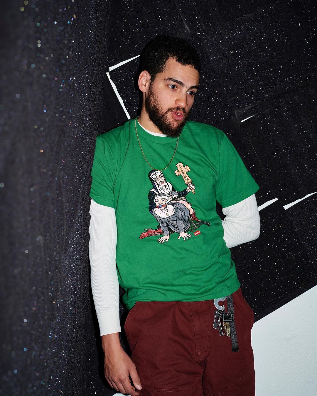 Man in green tee shirt with cartoon graphic stands near an indoor textured wall with keys hanging from his maroon pants. 