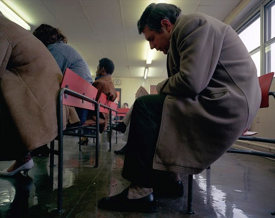 Several people are sitting in a waiting room with red chairs, captured in a medium shot showing men and women in coats indoors.
