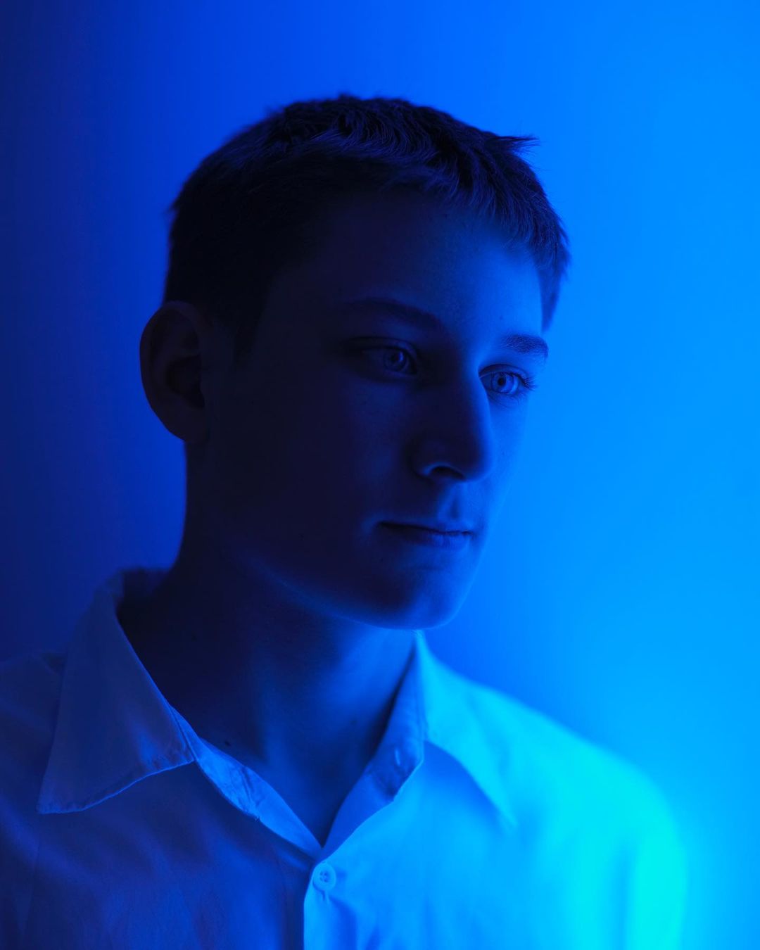 Portrait of a young adult male in a collared shirt, illuminated by blue lighting, casting shadows and highlighting the subject's features.