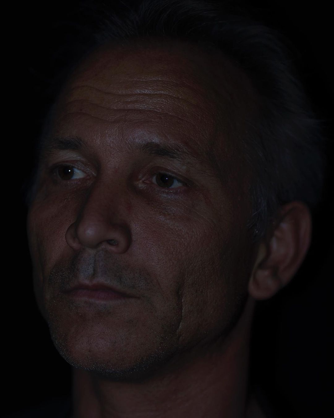 Headshot portrait of an adult male with short gray hair, fair skin, and a serious expression against a dark background.