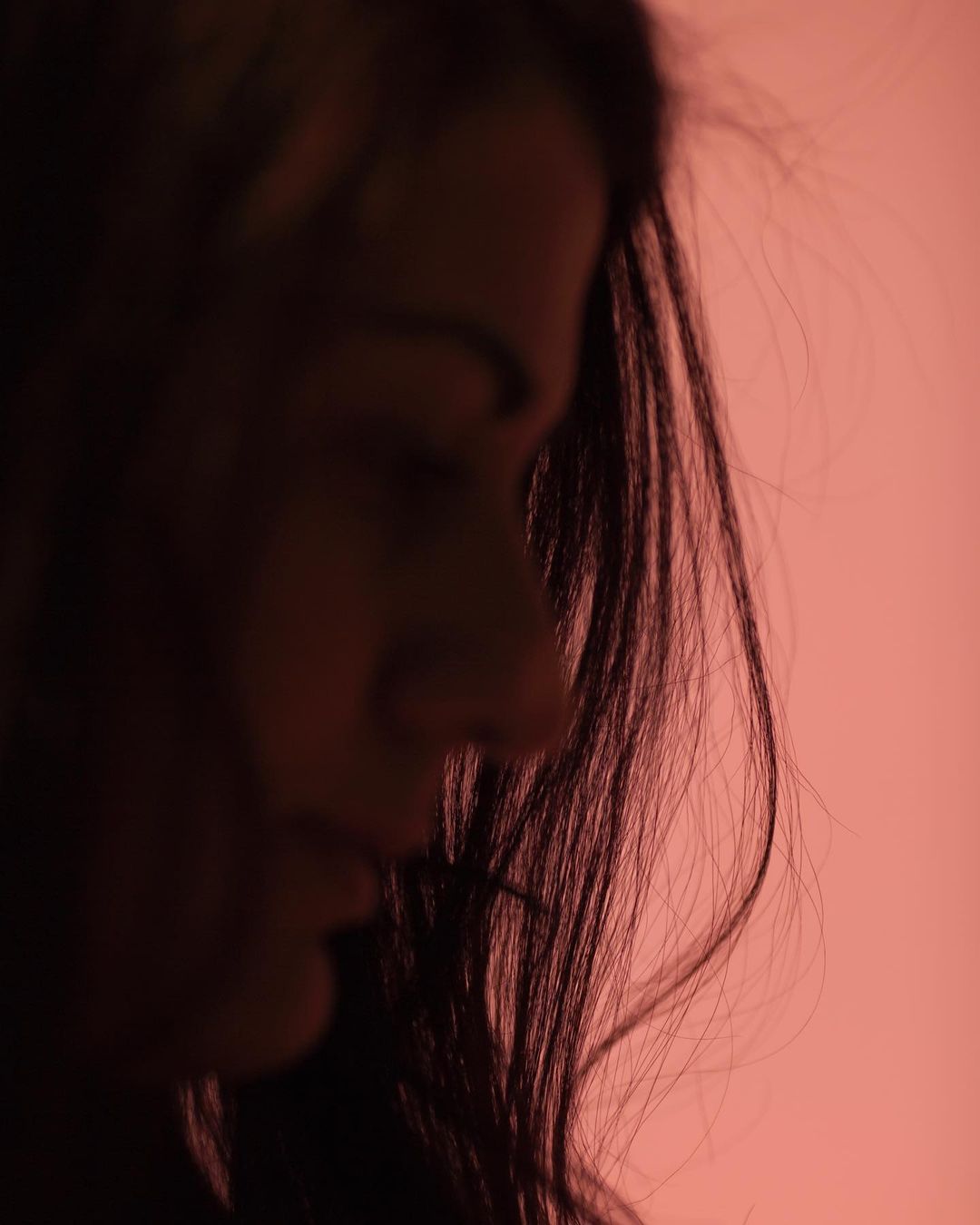 Close-up profile of a young adult female with long dark hair, under soft red lighting, against a pink background. Studio portrait.