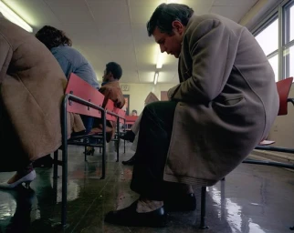 Man seated wearing a beige coat in a waiting area with red chairs and several other people in the background