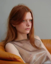 Portrait of a young woman with long red hair and a sheer top seated against a plain neutral background.