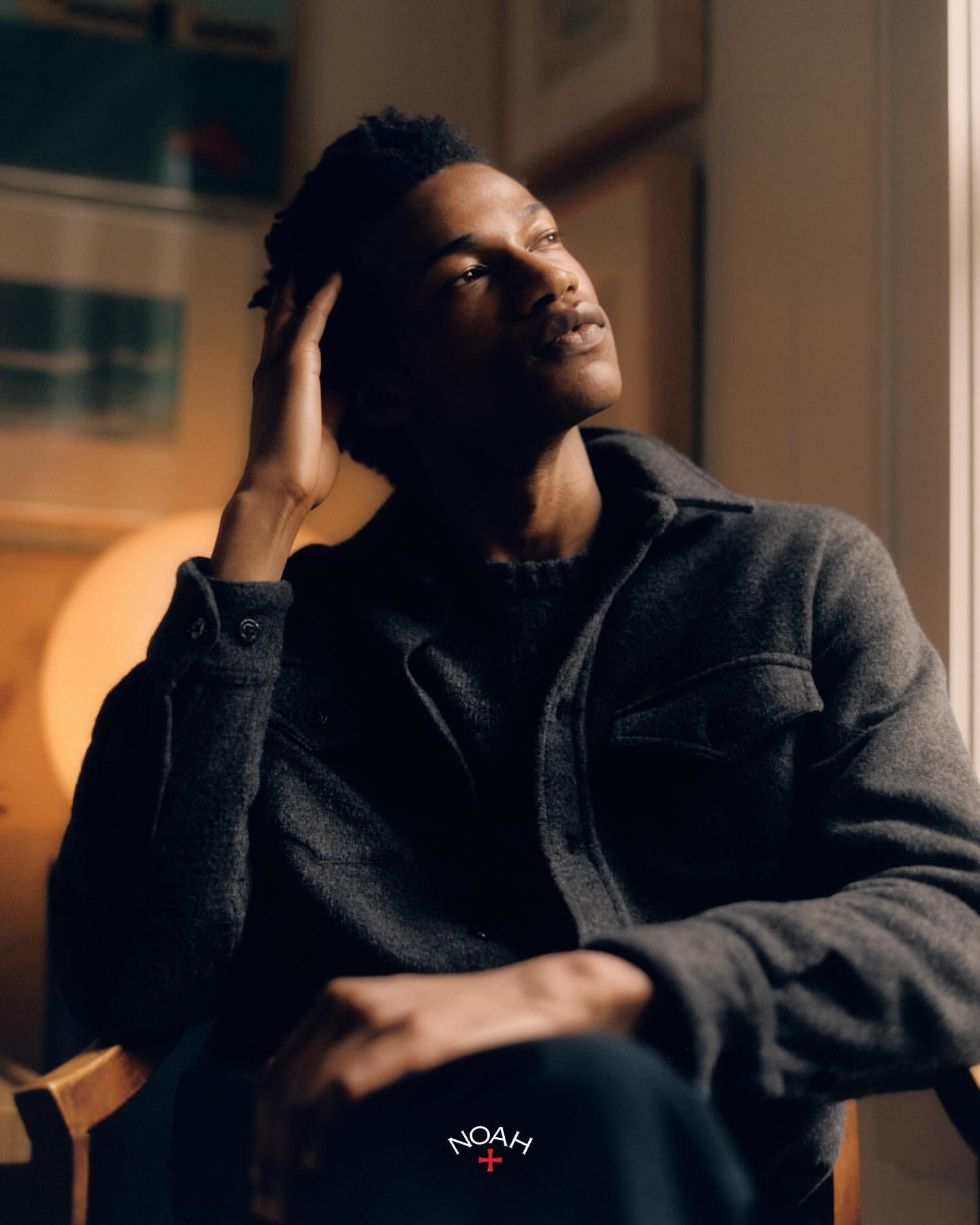 Portrait of a Black man in a collared jacket sitting indoors with a thoughtful expression, soft lighting, and muted neutral colors.
