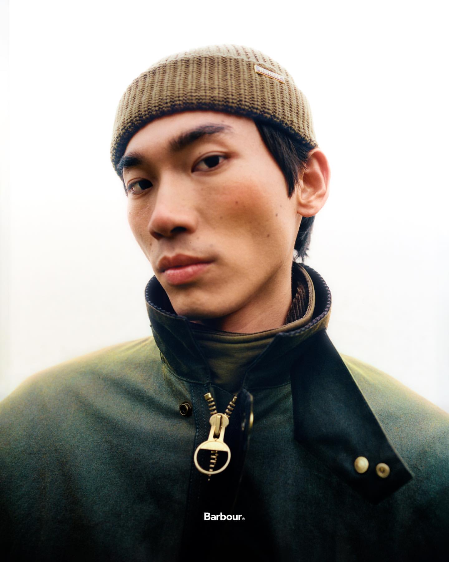 Portrait of a young Asian man wearing a beanie and Barbour jacket. Close-up shot, eye-level perspective, against a bright, neutral background.