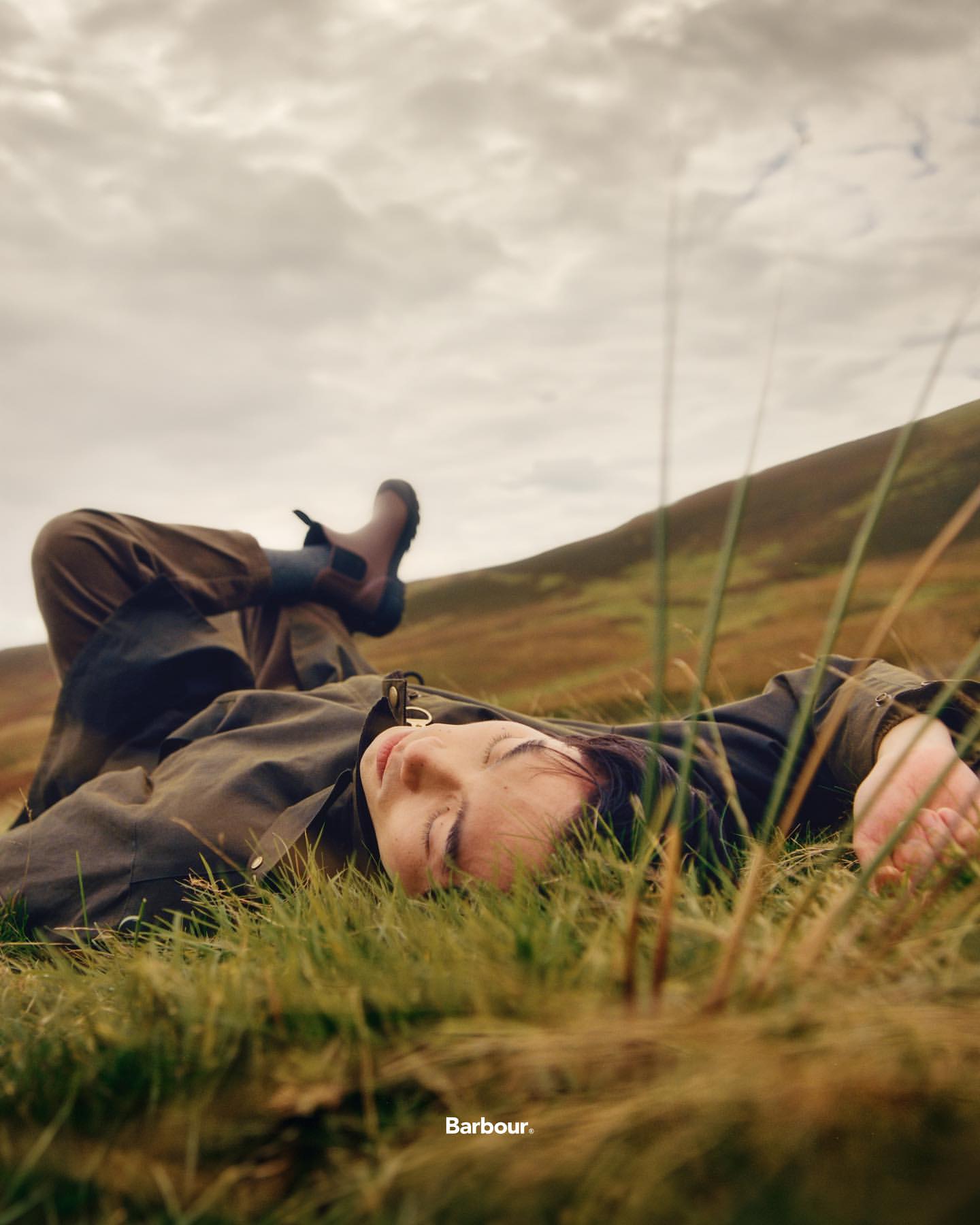 Woman lying on a grassy hill wearing Barbour brand jacket and boots with a cloudy sky in the background, fashion advertising shot.