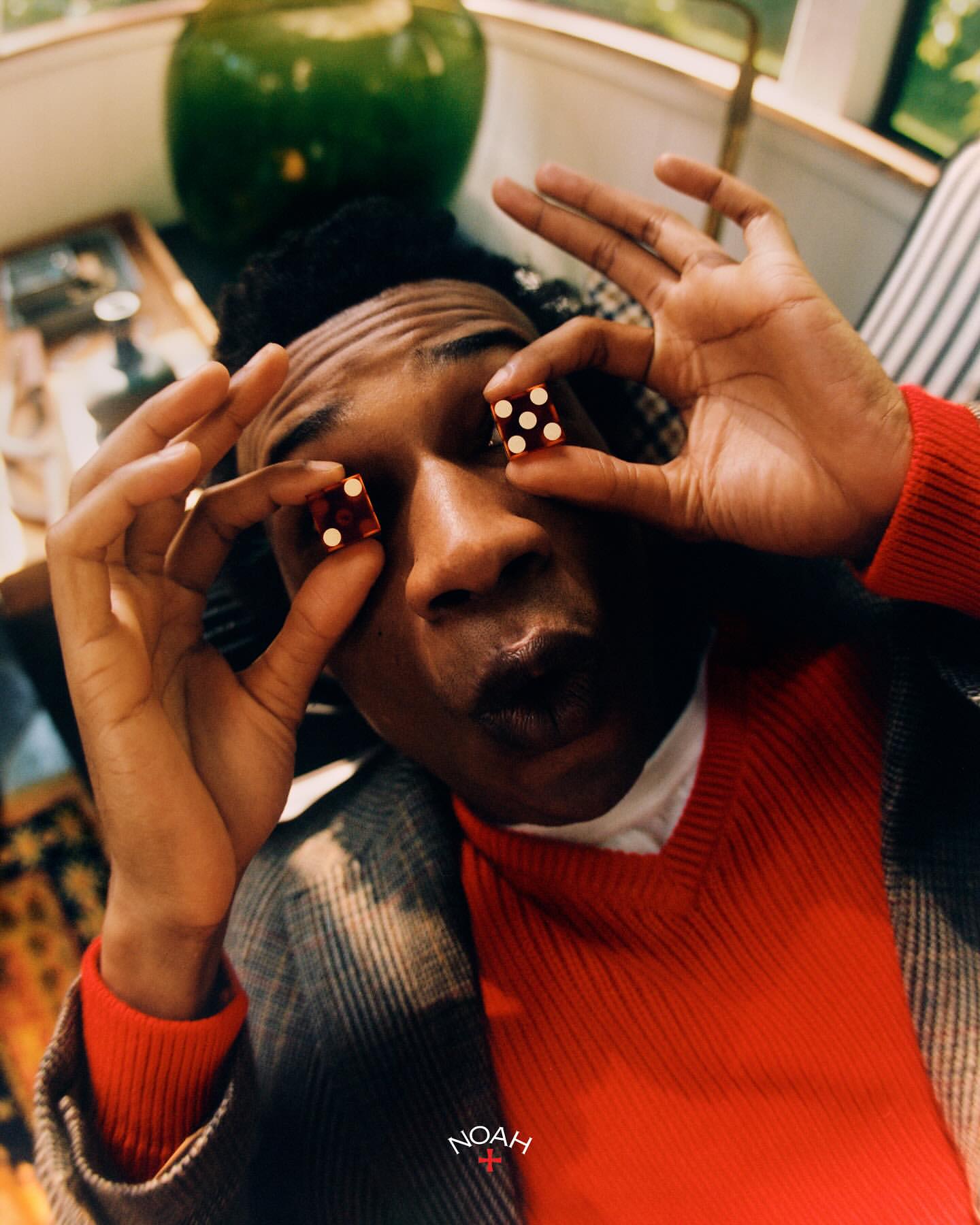 Portrait of a smiling Black man indoors holding red dice over his eyes, wearing a red shirt and plaid jacket.
