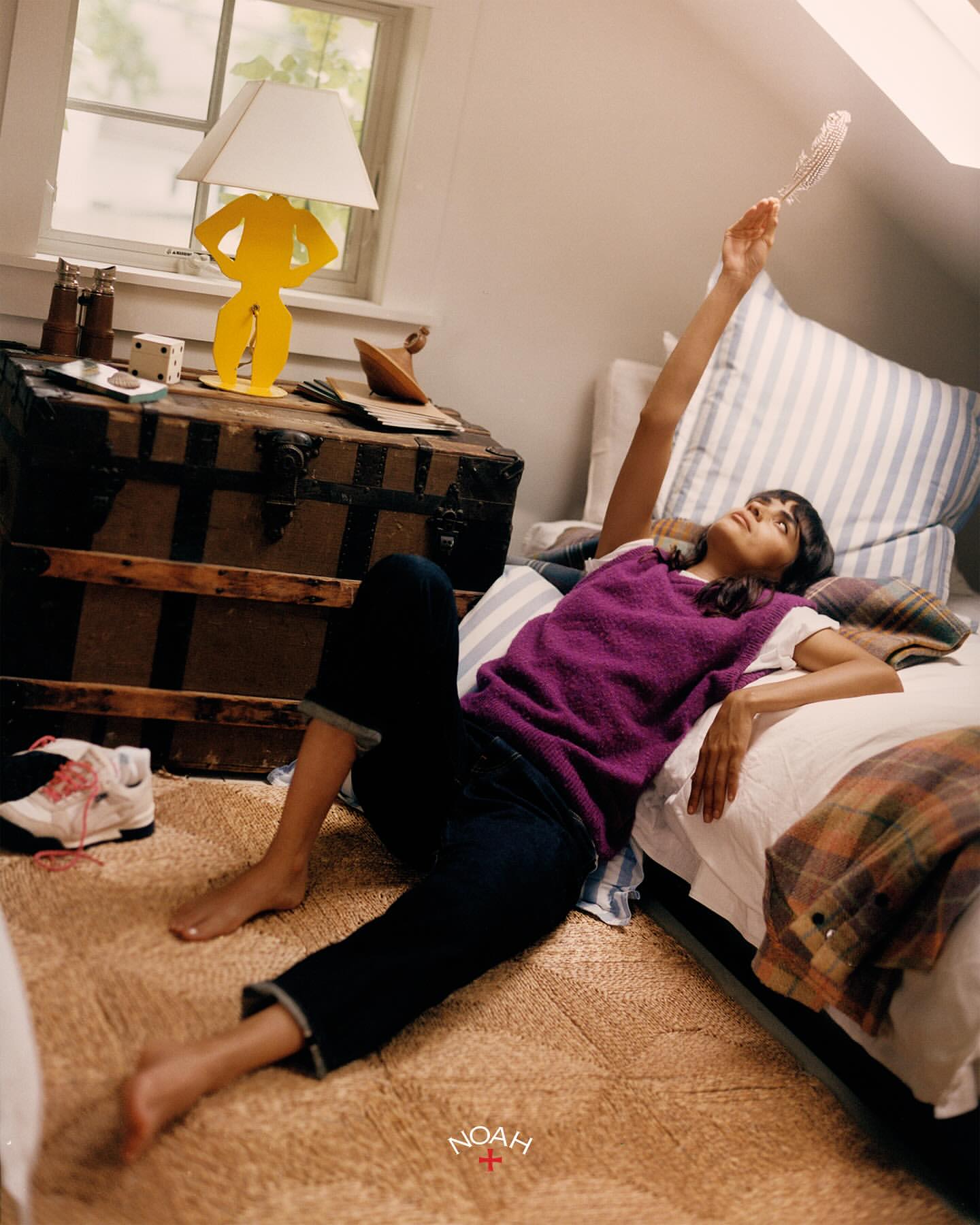 A young woman lounges in a sunlit bedroom, casually dressed, next to a bed and vintage trunk with a quirky yellow lamp and homey decor.
