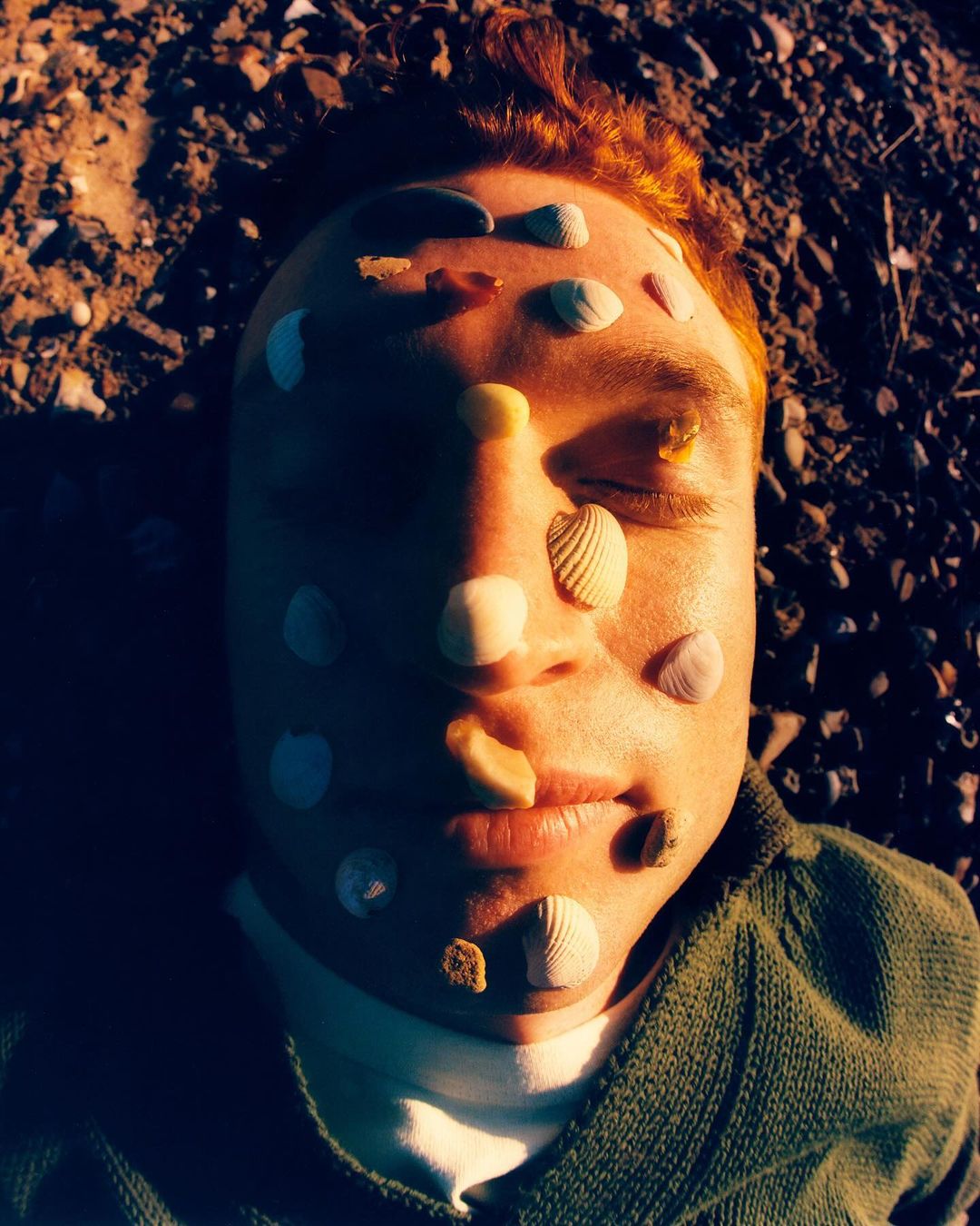 Close-up of a man lying on the ground with seashells on his face, illuminated by natural light and set against a blurred background.