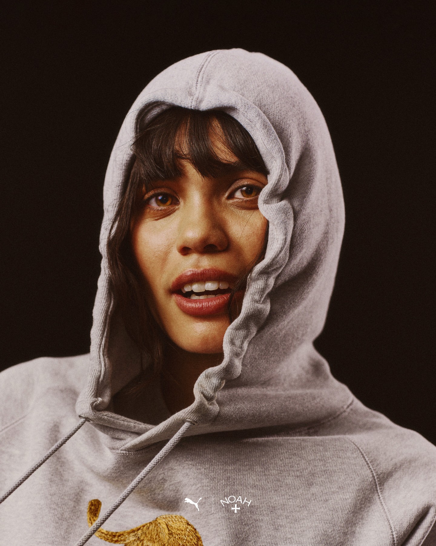 Close-up studio portrait of a young adult female wearing a grey hooded sweatshirt with a graphic on the front against a black background.