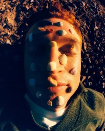 Close-up portrait of a man with red hair lying down with small seashells and stones on his face.