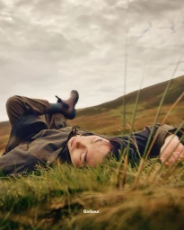 Person lying on a grassy hillside wearing a dark wax jacket and boots under an overcast sky, captured by Paul Hempstead.