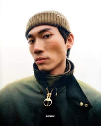 Man in olive green beanie and matching wax jacket looking down at the camera against a bright background