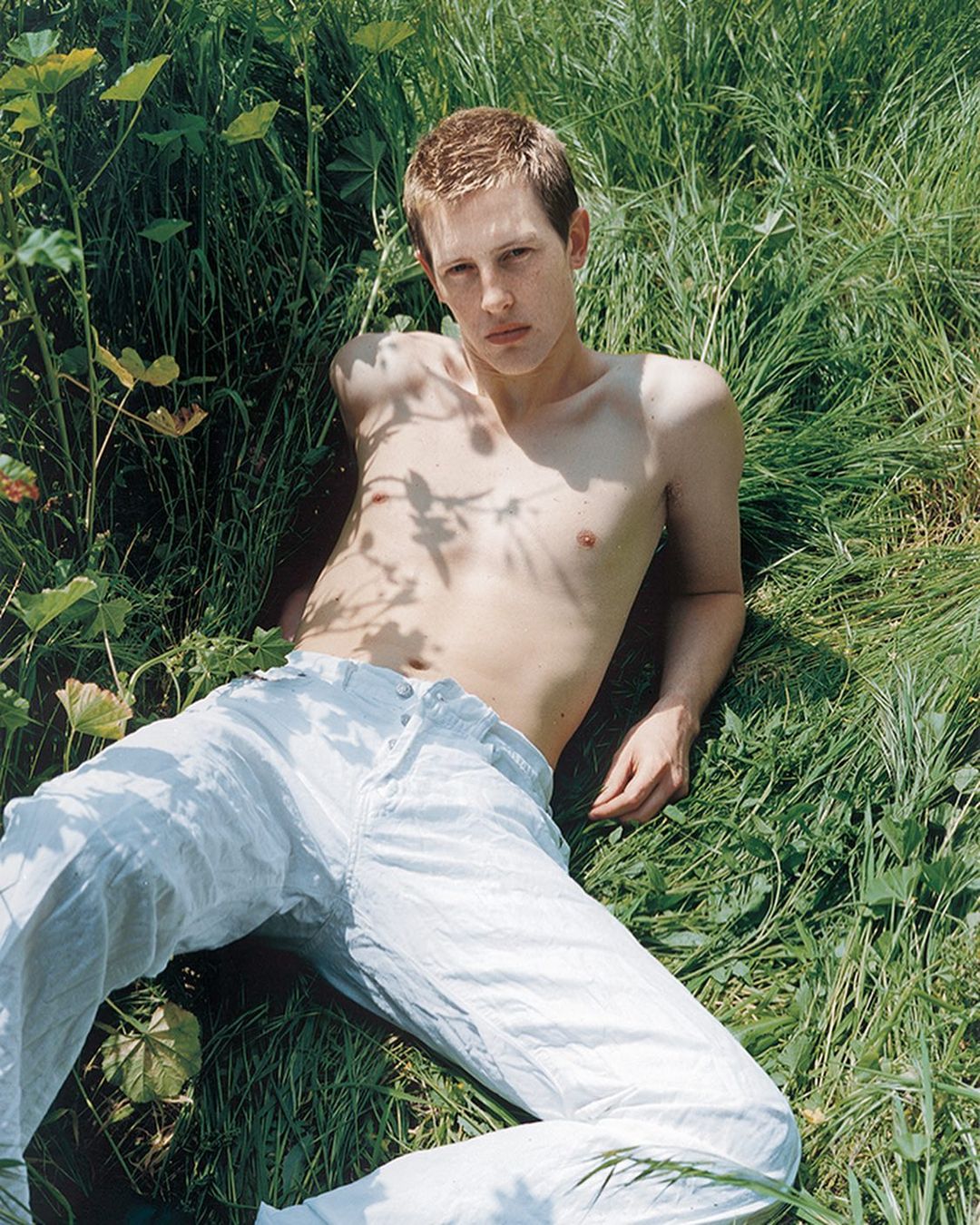 Young adult lies shirtless in grass outdoors. Casual fashion and nature combine in this eye-level shot with natural lighting.