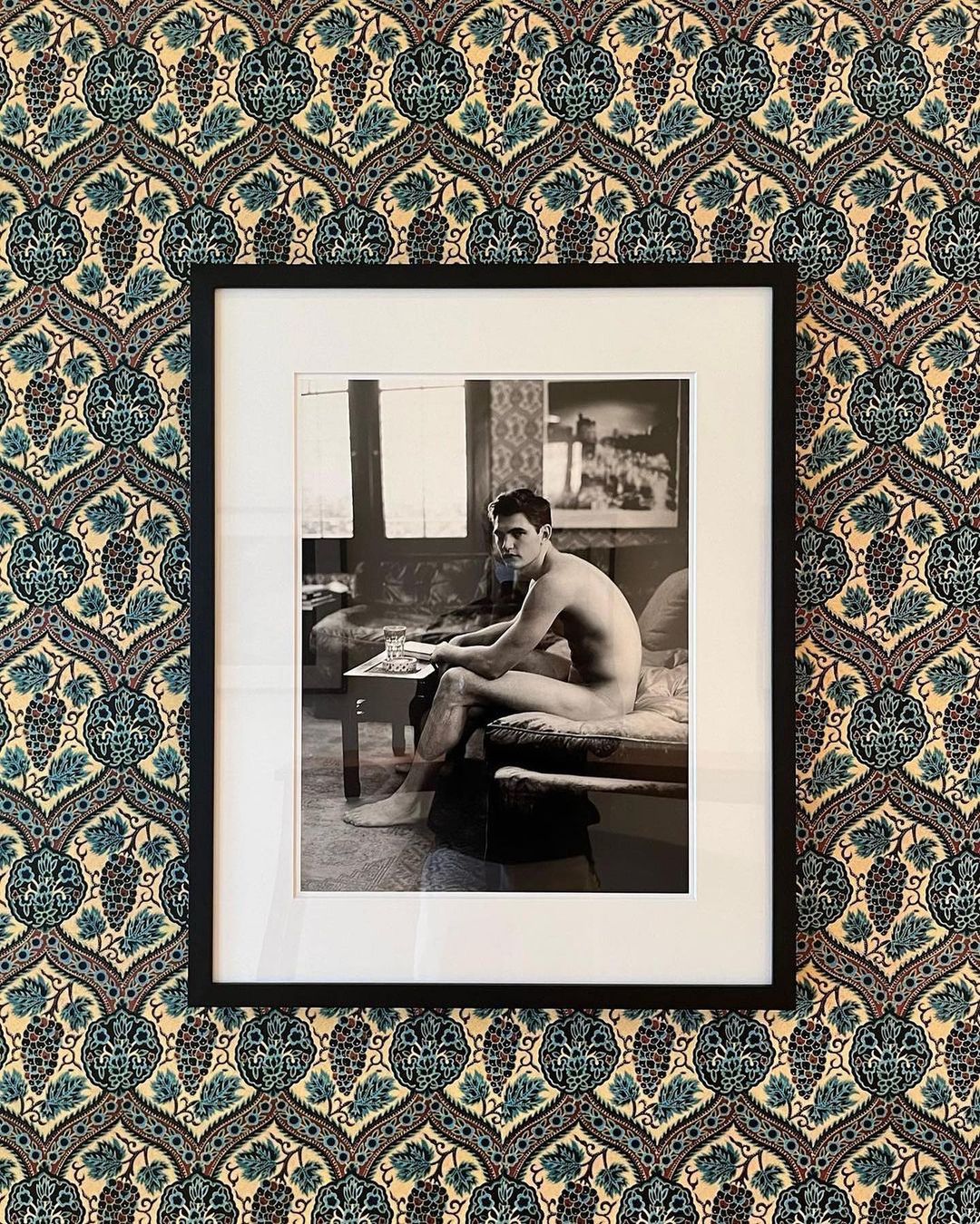 Black and white framed photo of a nude man sitting on a couch, displayed against a patterned blue and beige wallpaper background.