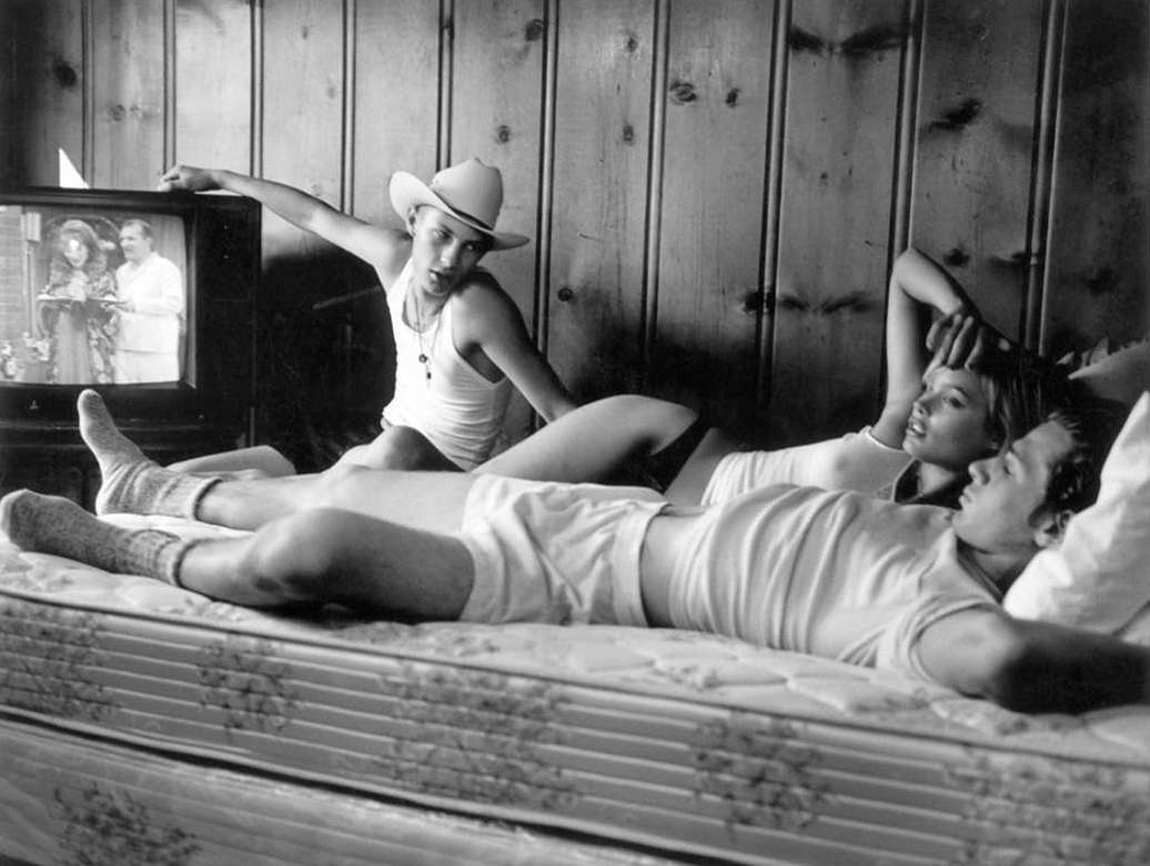 Black and white photo of three people lounging on a bed watching an old television with a wood paneled wall as a backdrop.