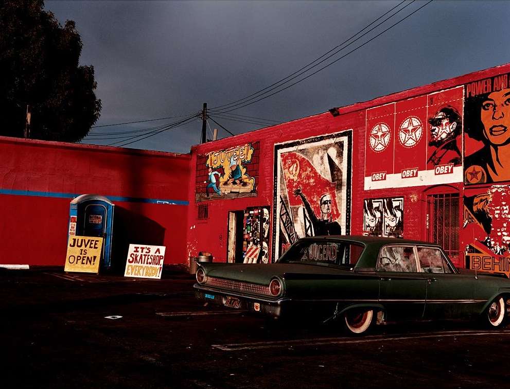 Vintage dark green car parked near a red brick building with advertising signs and posters, captured at dusk with dark shadows.