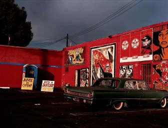 Vintage dark green sedan parked in front of a red building covered with bold street art posters and signs.