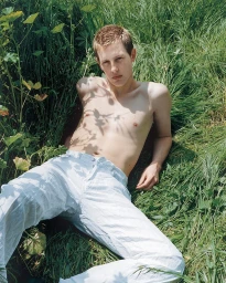 Shirtless young man wearing white trousers lying in tall green grass outdoors