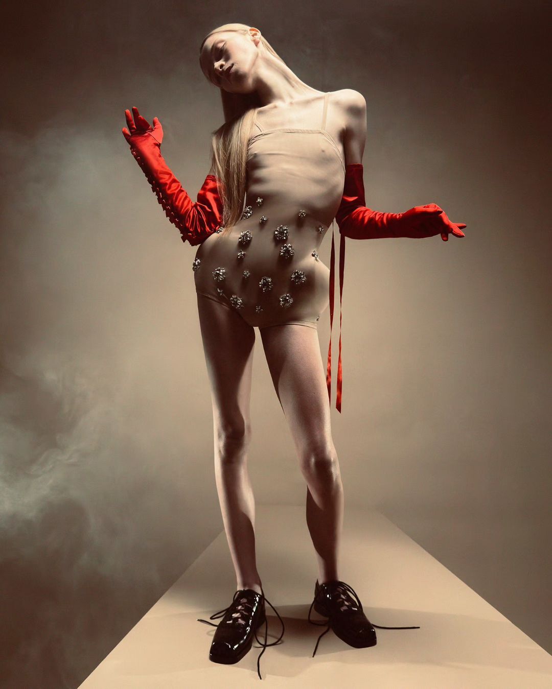Full-length shot of a slender woman in a beaded bodysuit, red gloves, and platform shoes, on a beige surface with a smoky background.