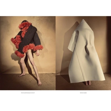 Two figures in avant-garde fashion garments pose in a neutral setting with one figure in a dark furry coat and the other in white garment.