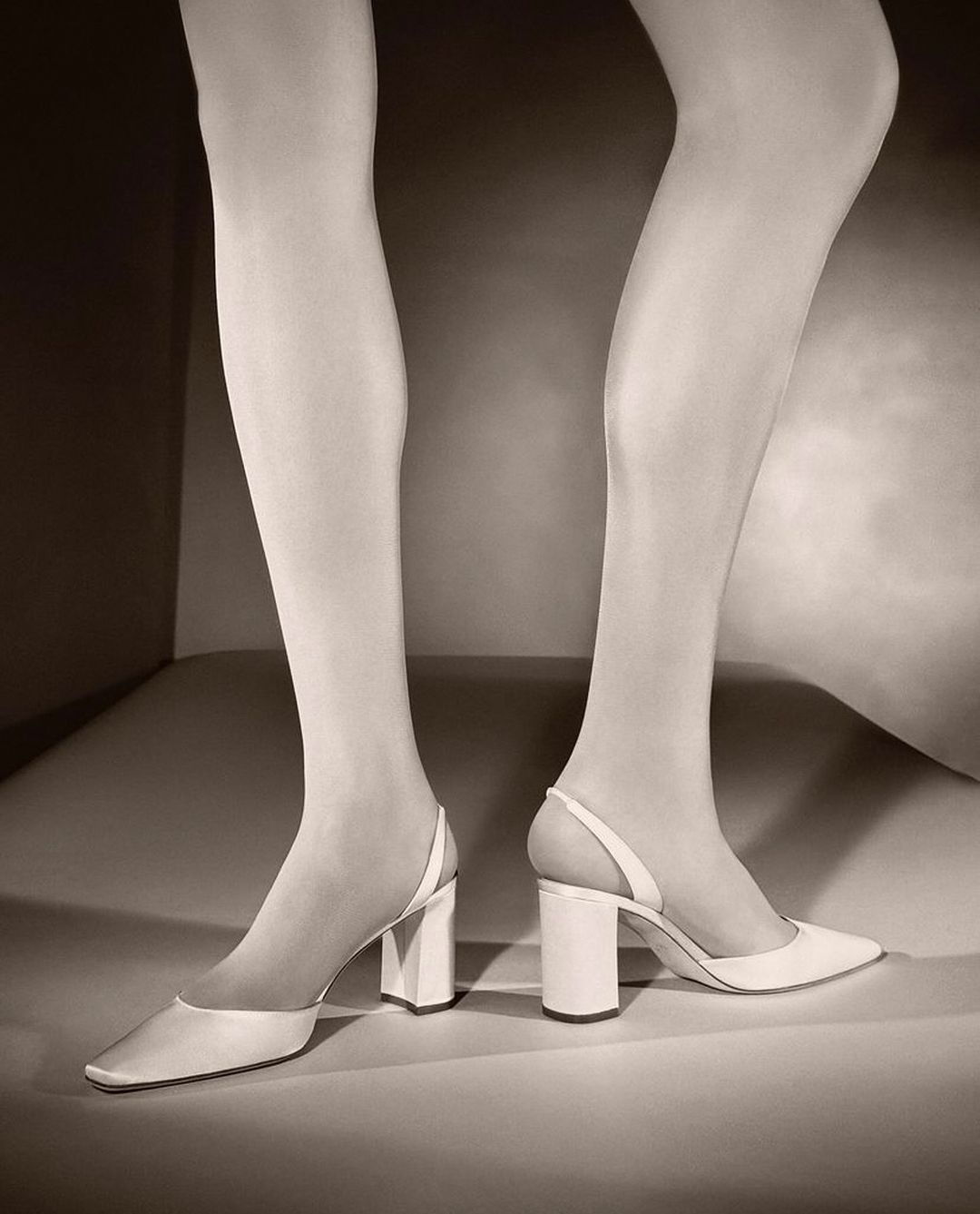 Close-up fashion shot of a woman's legs wearing white tights and pointed-toe white heels in a minimalist studio setting.