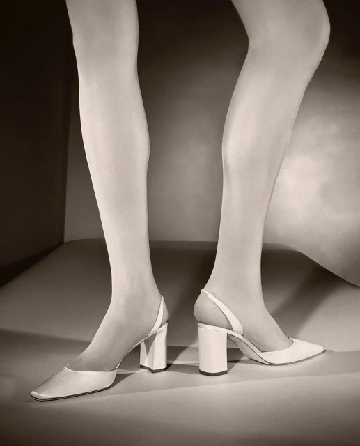 Close-up fashion shot of a woman's legs wearing white tights and pointed-toe white heels in a minimalist studio setting.