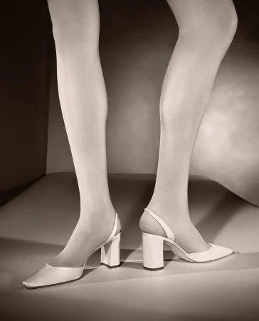 Close-up fashion shot of a woman's legs wearing white tights and pointed-toe white heels in a minimalist studio setting.