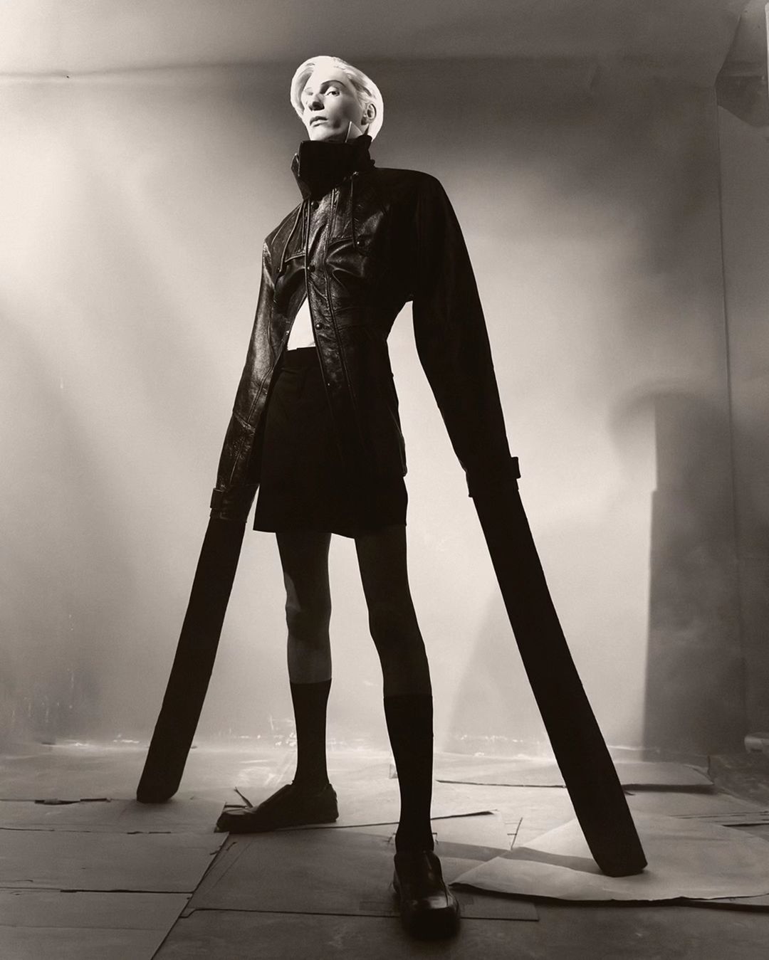 Monochrome studio shot of a slender adult female fashion model in black coature, wearing black hosiery and pumps. She is standing and looking up.