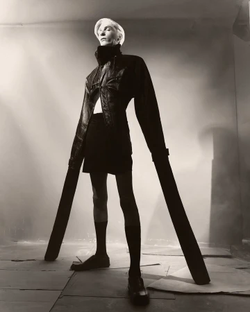 Monochrome studio shot of a slender adult female fashion model in black coature, wearing black hosiery and pumps. She is standing and looking up.