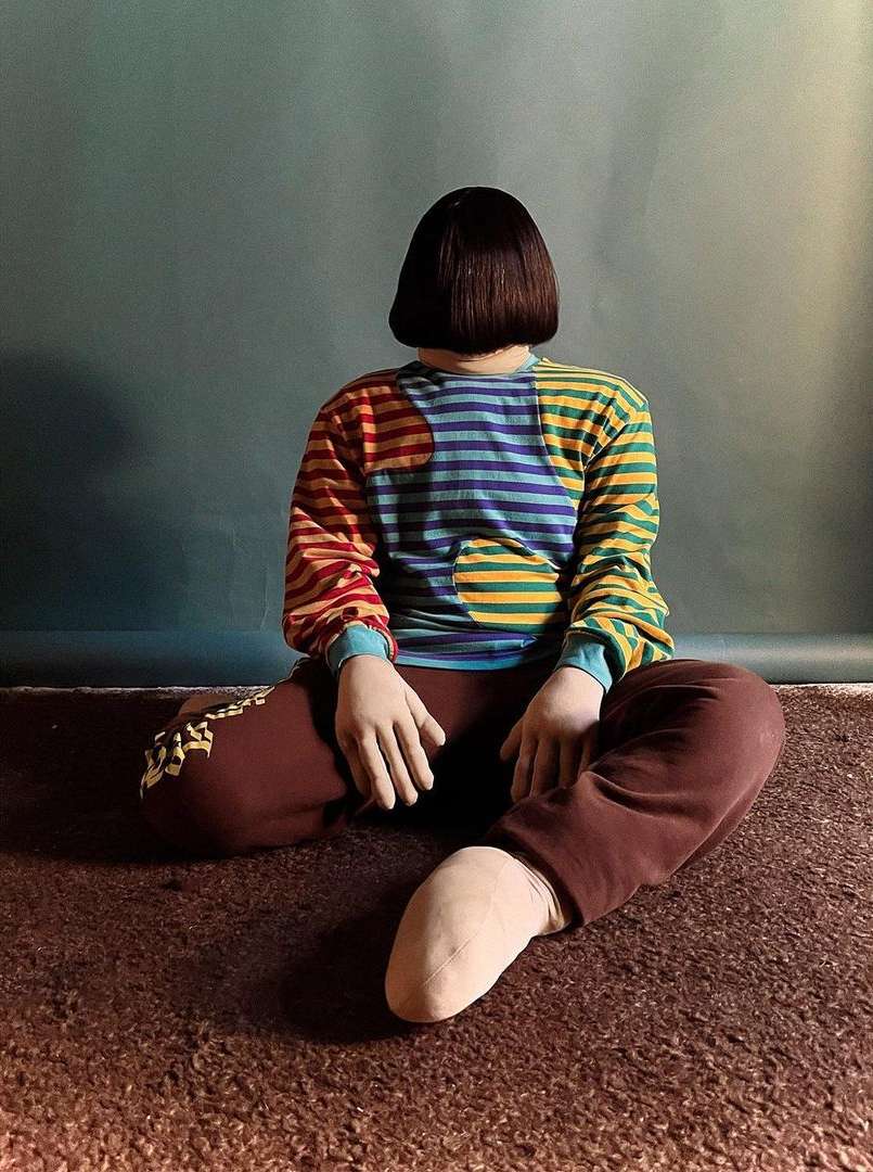 Person sits cross-legged on a shag rug, facing away with colorful striped top, and short brunette bob hairstyle against a teal wall.