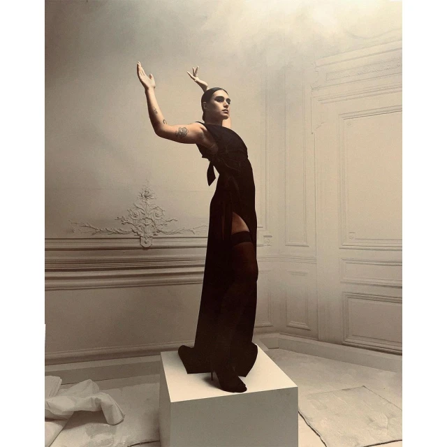 Adult female fashion model in a dark dress and boots poses with raised arms on a white cube in a white room with wall molding.