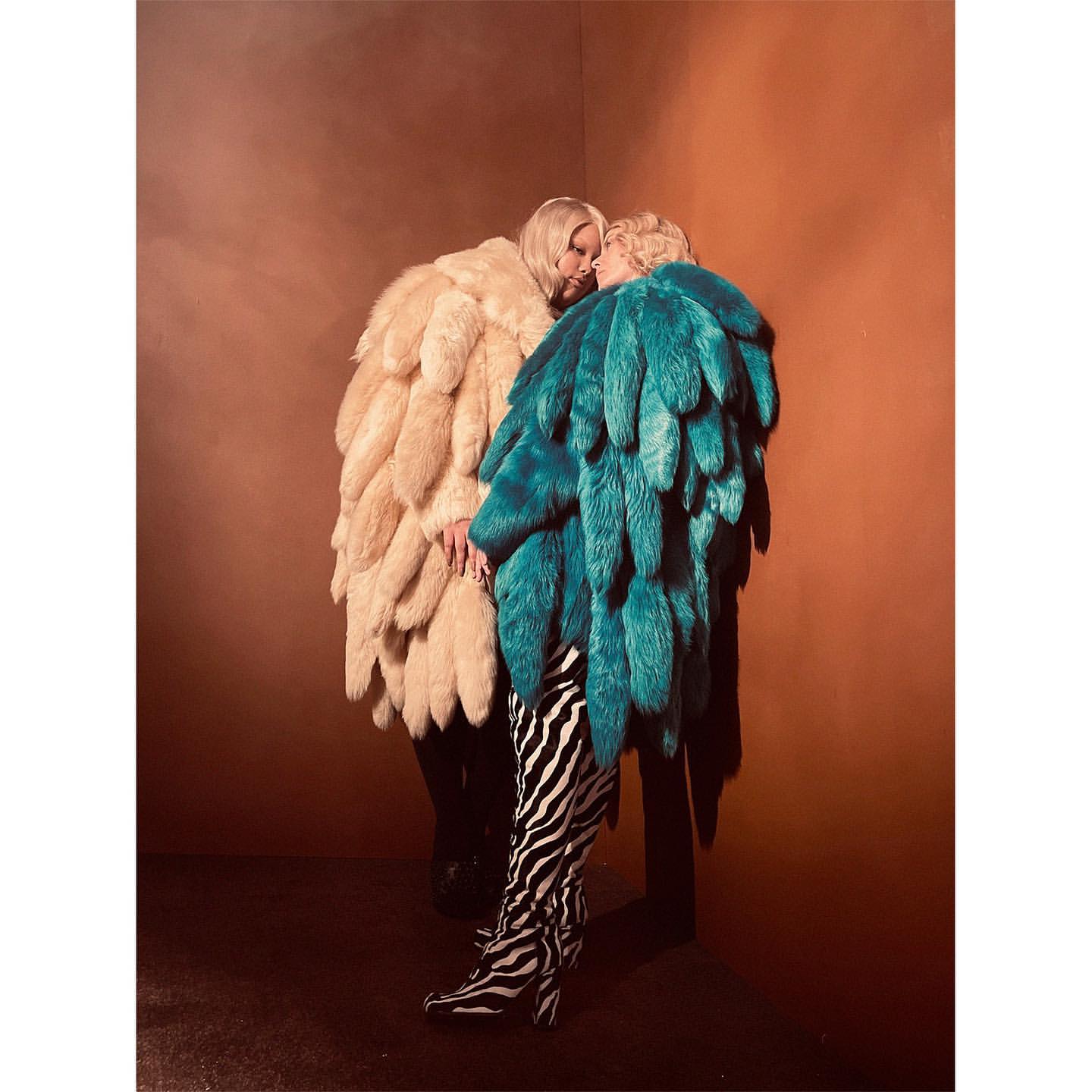Two people in large feather coats stand close together in a corner with an orange wall, one wearing zebra print boots.